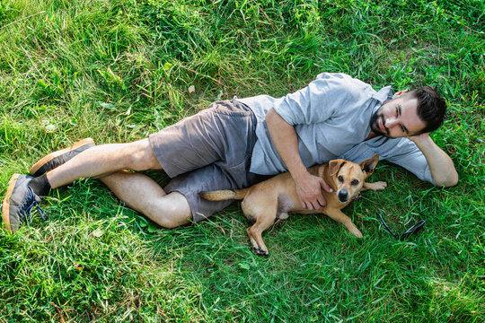 Smiling attractive European man is lying on grass and hugging his cute little dog.