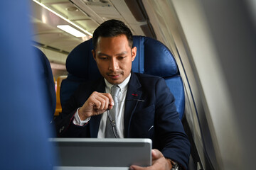 Asian businessman in suit using digital tablet while sitting comfortable seat in airplane cabin