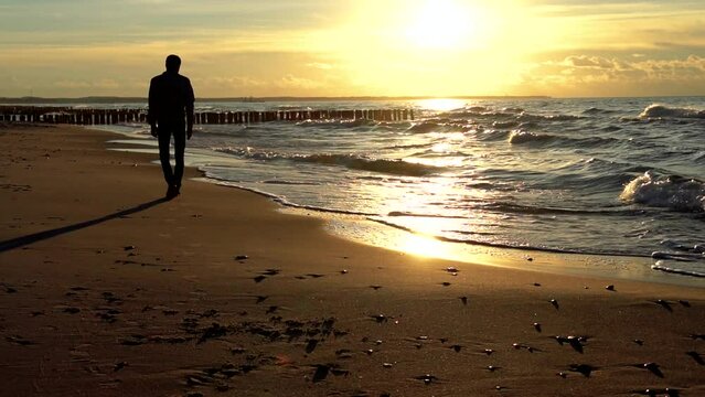 Man walking on a beach at golden sunset. Wavy Baltic sea with breakwater. Normal speed.