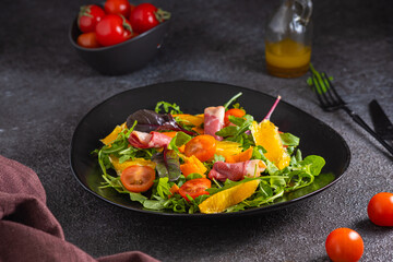 Fresh salad with arugula, cherry tomatoes, orange slices and smoked duck slices on a black plate on a dark concrete background. Served with olive oil dressing. Salad recipes.