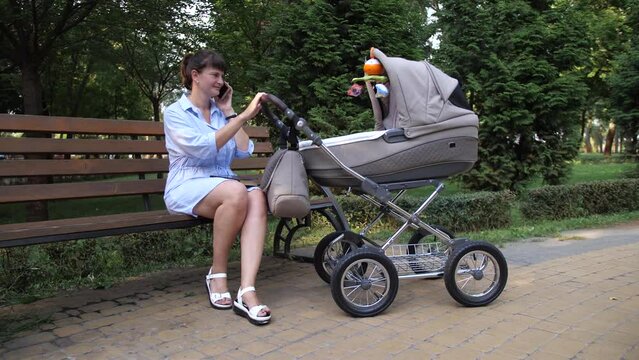 A young mother sits on a bench, shakes a stroller talking on the phone. The camera is in motion. 4K Slow Mo