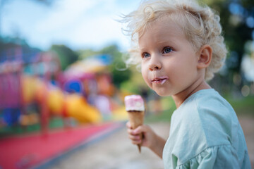 The child eats ice cream in the park in the summer on the background of the playground. Close-up portrait of pink ice cream curly-haired child girl blonde.