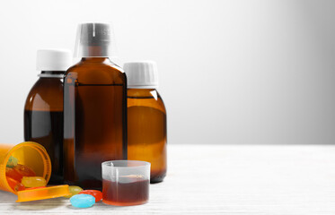 Bottles of syrup, measuring cup, lozenges and spoon on white wooden table against light grey...