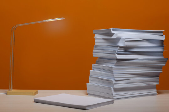 Stack Of Paper Sheets And Lamp On White Wooden Table Near Orange Wall