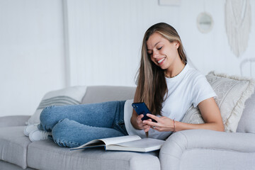 Smiley beautiful caucasian woman in white t-shirt, blue jeans laying on sofa with book holds phone smiles makes video call toothy smiles relaxing home. Attractive Italian female talks with boyfriend.