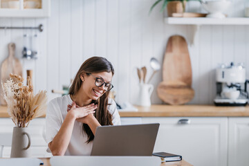 Adorable female student in white t-shirt sitting at kitchen table with laptop puts hands on chest feels gratitude after sistant lesson. Remote education. Pretty Italian woman makes video call at home.
