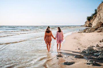 Two teen caucasian barefooted girls from behind wearing airy dresses holding their hands and walking along sandy coast with white rock in sunny summer day. Sisterhood and friendship