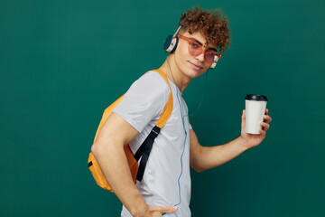 a man in gray clothes is listening to music in headphones with bright glasses and a backpack on his back, holding a cardboard glass for hot drinks in his hand, standing sideways to the camera