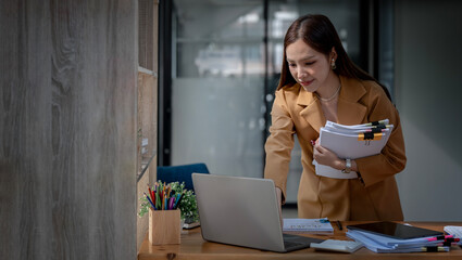 Attractive businesswoman carrying a stack of documents and working on laptop computer, standing at...