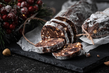 Chocolate salami filled with almonds, hazelnuts and raisins on the black background. Italian sweet sausage shaped dessert sprinkled with powdered sugar. Christmas and New Year festive pastry