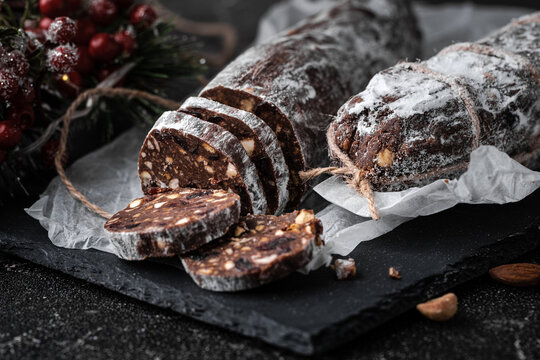 Chocolate salami filled with almonds, hazelnuts and raisins on the black background. Italian sweet sausage shaped dessert sprinkled with powdered sugar. Christmas and New Year festive pastry