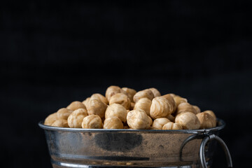 A pile of peeled hazelnuts in a metal bowl on the black background. Healthy nutrition concept. Traditional rustic agricultural background. Copy space for a free text. Macro shot