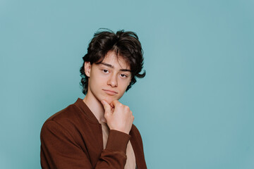 Thoughtful handsome teenager in brown jacket touching chin makes choice looks at camera stands against turquoise background. Confident Italian boy posing at studio. Male model.