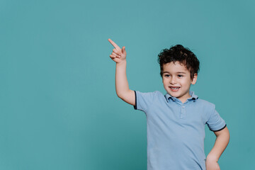 Nice little boy in blue polo pointing at empty space standing against turquoise studio backdrop. Handsome Spanish kid smiles points by index finger. Cute toddler, mockup, discount, promo, sale.