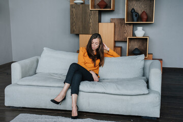 Obraz premium Depressed brunette hispanic woman in orange blouse sitting on cozy sofa with upset facial expression, tired frustrated girl after divorce failure mistake. Perplexed lady sitting on sofa at home.