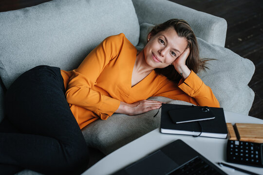 Pretty Hispanic Brunette Young Woman In Orange Blouse And Black Pants Laying On Cozy Couch Looks At Camera Relaxing Of Remote Work. Beautiful Caucasian Female Student After Distant Lesson Via Internet