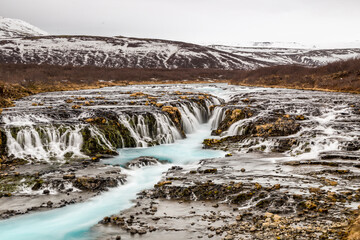 Brúarárfoss, cascata ampia con poco salto, acqua azzurra, con sfondo di montagne innevate e bosco