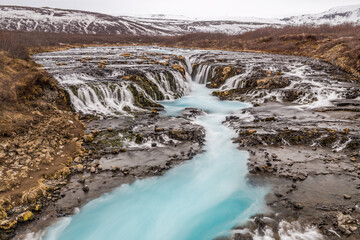 Brúarárfoss, cascata ampia con poco salto, acqua azzurra, con sfondo di montagne innevate e bosco