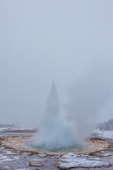 Geyser in eruzione durante una nevicata con cielo grigio e terreno marrone innevato