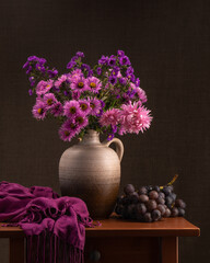 Many pink, purple flowers in a gray clay vase, jug on a wooden table. Nearby lies a dark gray shawl...