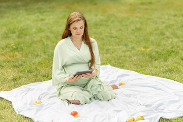 Young pretty pregnant woman holding, reading something on tablet,gadget,resting in park,sitting on cover,blanket in park,meadow. Healthy walk outdoors. Preparation for childbirth. Horizontal