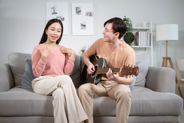 Happy young Asian couple singing a song playing  guitar on sofa in living room at home.