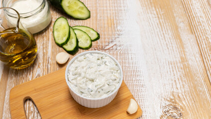 Traditional greek yogurt sauce Tzatziki,dressing in white bowl, made of grated cucumbers, sour cream yogurt,olive oil,garlic,herbs with ingredients on wooden table. Copy space, horizontal