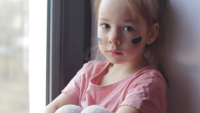 a sad little girl with a Ukrainian flag painted on her face is sitting looking out the window