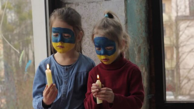 two little girls with the flag of Ukraine painted on their faces are sitting near the window with candles in their hands. children and the war in Ukraine.