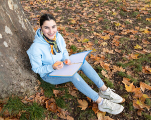 A student sits under a tree with a blue folder in her hands and smiles, Austria, Salzburg