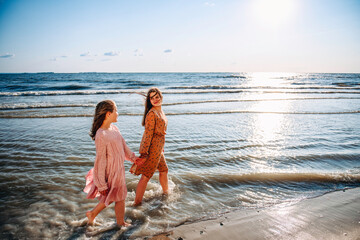 Two happy laughing teen caucasian barefooted girls wearing airy dresses holding their hands and walking along the beach in sunny summer day. Sisterhood and friendship