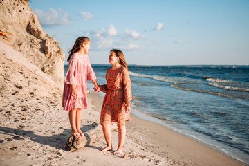 Two happy laughing teen caucasian barefooted girls wearing airy dresses holding their hands and walking along sandy coast with white rock in sunny summer day. Sisterhood and friendship