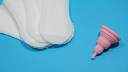 Copa menstrual rosa plegable y compresas desechables en un fondo azul. Transición a la...