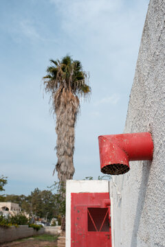 White wall with red pipe and red door. Palm tree and blue sky in the background. Creative image with trendy color 2023.