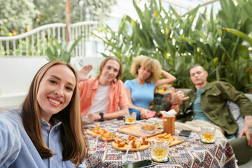Cheerful young woman taking selfie with group of her friends when they are enjoying dinner in outdoor cafe