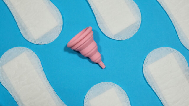 Copa menstrual rosa plegable entre compresas en un fondo azul. Menstruaci&oacute;n sostenible