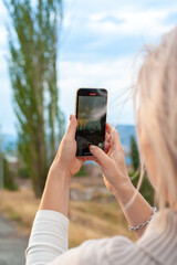 close-up of a young woman photographing a beautiful picturesque landscape at sunset using a mobile phone holding a smartphone with both hands. Looking behind