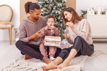 A happy young family is sitting on a knitted plaid, holding a wooden box with Christmas toys.