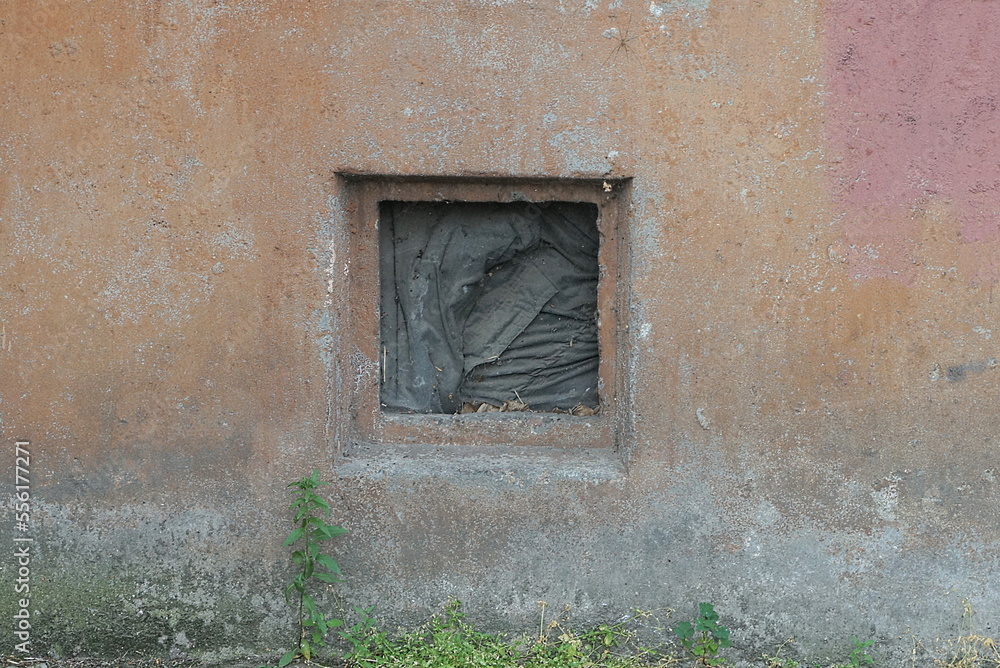 Sticker one small square old black window in the brown concrete foundation of the wall of the house on the street in the green grass