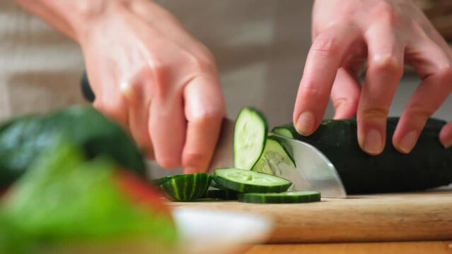 the cook cuts a spring salad of tomatoes, cucumbers, greens and mixes. Woman cutting cucumber for salad on wooden cutting board on kitchen table with cucumbers, tomatoes branch and scallion around.