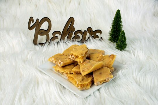 Peanut brittle on a white plate on white fur with miniature pine trees and believe Christmas sign 