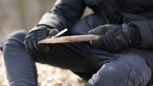 Man using knife to sharpen tree branch preparing camp in woods on cold autumn day