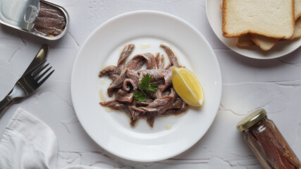 Plate with canned anchovy fillets, lemon wedge and pieces of toast on white textured table, flat lay