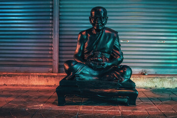 Black Buddha statue in the streets of Bangkok at night in front of a closed roller gate. Cinematic look of red and blue light.