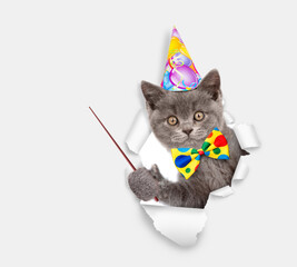 Cute cat wearing party cap and tie bow looking through a hole in white paper and pointing away on empty space