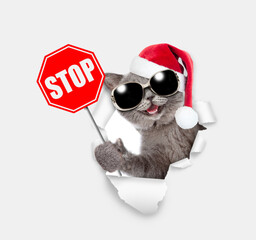 Happy kitten wearing sunglasses and red santa hat looking through a hole in white paper  and shows stop sign
