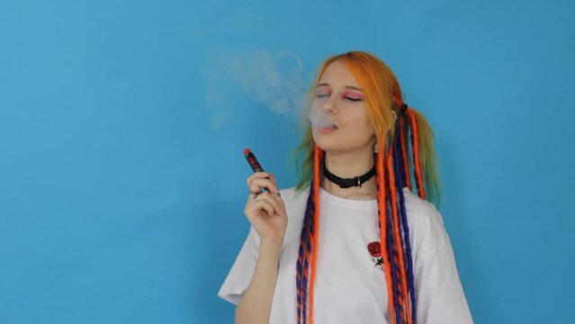 A modern hipster girl with colored hair smokes a vape on a blue background, A fashionable new vaping device, smoke e-liquid instead of nicotine cigarettes