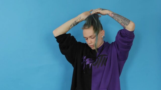 A stylish LGBT guy tucks his hair into a hair scrunchie and smokes an electronic cigarette while sexually releasing smoke on a blue background in the studio.