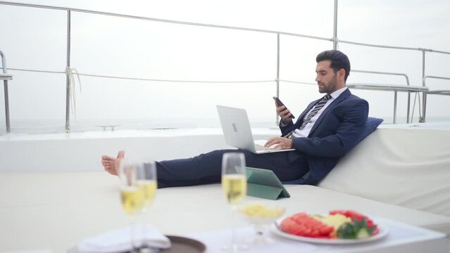 Caucasian businessman working on laptop computer and talking on mobile phone for global corporate business while travel on luxury private catamaran boat yacht sailing in the ocean on summer vacation.