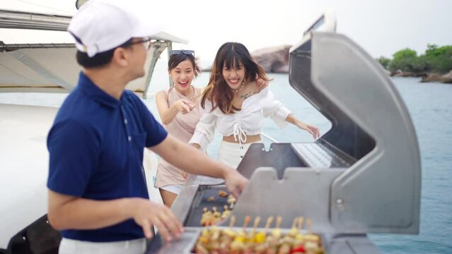 4K Asian man chef cooking grilled barbecue and seafood on stove for serving to woman passenger tourist travel on luxury private catamaran boat yacht sailing in the ocean on summer holiday vacation.